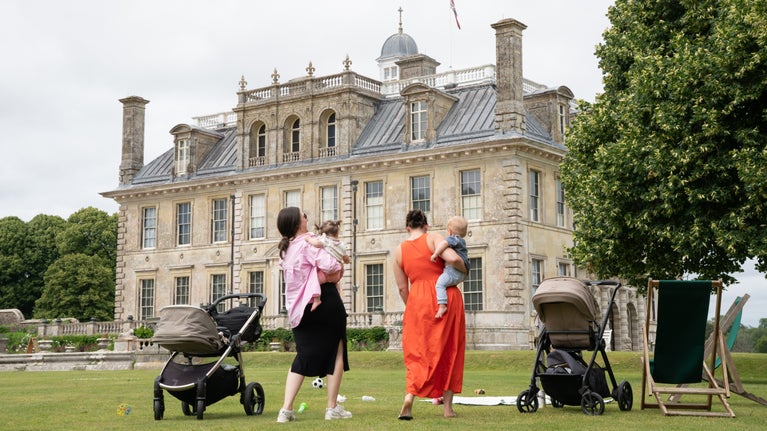 In front of a stone mansion two women hold young children. There are buggies either side